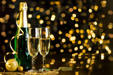 Two glasses and green bottle with champagne, gold decor, balls, serpentine are on table. Festive decorative garland, yellow light bulbs, lanterns are shining on background. New year, christmas mood.