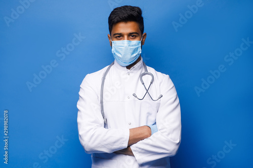 A indian surgeon standing setting his gloves to start the surgery, wearing a mask and a blue uniform, isolated on a blue background.