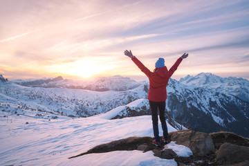 Happy young woman in snowy mountains at sunset in winter. Beautiful slim girl on the mountain peak with raised up arms, snow covered rocks and colorful sky with clouds. Travel in Dolomites. Tourism