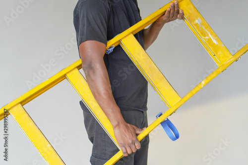 African electrician holds a yellow wooden ladder. The builder bears a wooden step-ladder on a gray background. Factory worker with a yellow wooden staircase. photo