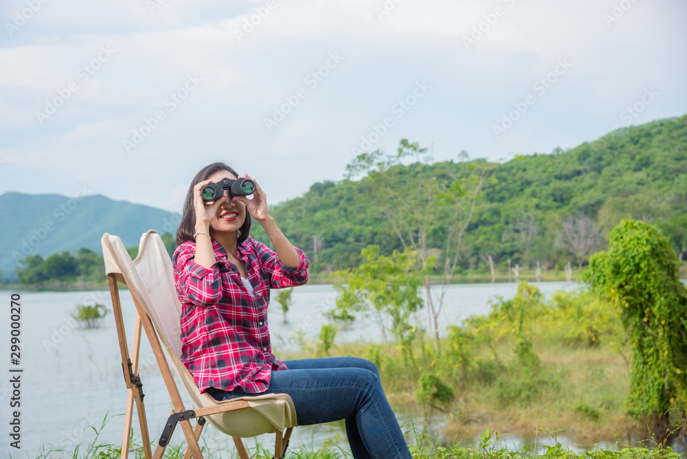 Beautiful Asian woman using binoculars to see the birds between camping at lake side camping site.
