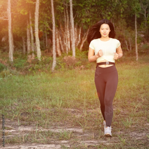 Young Asian woman wears sport black leggings runs on rural road in tropical rain forest in the morning with warm sunshine. Female runner practice trail running in forest. Health care and Lifestyle.