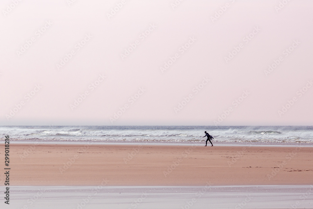 Lone surfer, carrying a surfboard, on a beach in Cornwall, UK