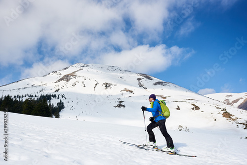 Male hiker, tourist in colorful clothing with backpack trekking on skis up snowy hill on background of bright blue sky and beautiful mountain on frosty sunny day. Winter active vacations concept.