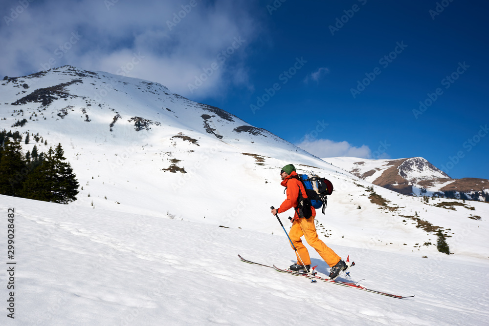 Skier tourist backpacker walking on skis up steep hill on background of bright blue sky and beautiful mountains ridges. Winter vacations, active lifestyle, skiing and trekking concept. Skier tourist backpacker walking on skis up steep hill on background of bright blue sky and beautiful mountains ridges. Winter vacations, active lifestyle, skiing and trekking concept.