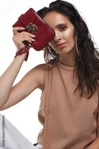 Cropped medium shot of a young dark-haired lady in a beige blouse showing a cherry-coloured haircell leather clutch with a long handle and a flap fastening with a golden case clasp photo