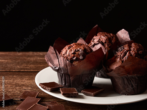 Still life with brown muffins with cocoa in a wrapping of baking paper, pieces of chocolate on a white porcelain plate and old wooden table top. Black background