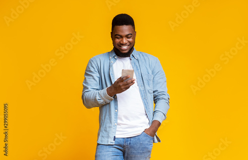 Cheerful black guy reading message on smartphone over yellow background