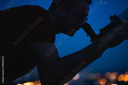 Astronomer with a telescope watching at the stars and Moon with blurred city lights in the background.