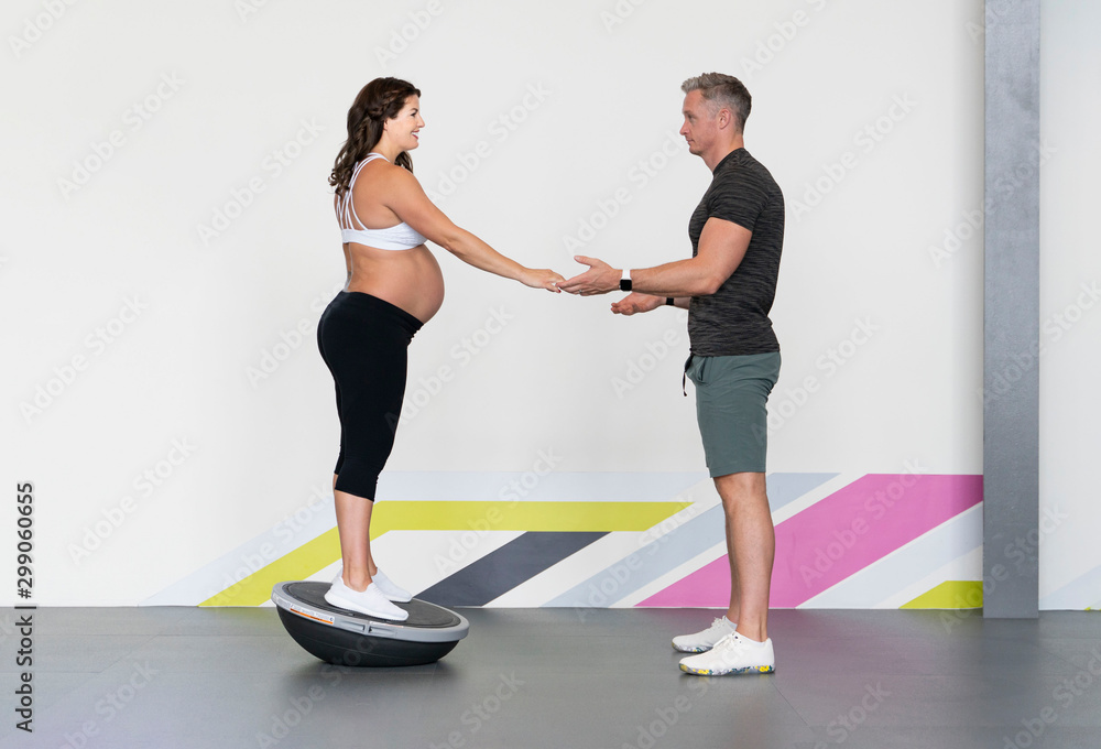 Caucasian pregnant woman in her third trimester doing bodyweight balance exercises with the help of her personal trainer in a gym wearing sports or fitness clothing Caucasian pregnant woman in her third trimester doing bodyweight balance exercises with the help of her personal trainer in a gym wearing sports or fitness clothing