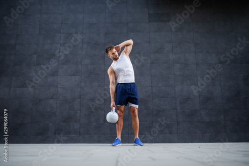 Full length of handsome strong muscular bearded Caucasian man in shorts and t-shirt posing with kettle bell while standing outdoors in front of gray wall.