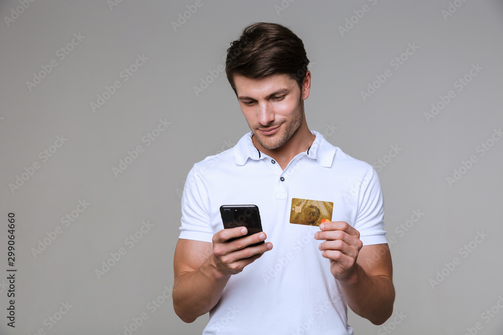 Concentrated pleased man holding debit card using mobile phone. Concentrated pleased man holding debit card using mobile phone.