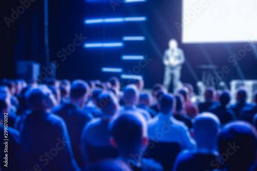 Crowd of seated business people in the background of the presenter on stage. Defocused blurred picture for background