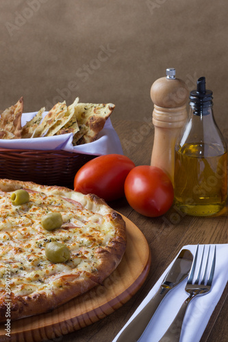 Gratin Loin Pizza. Mozzarella, Canadian Sirloin, Creamy Curd and Green Olives. Accompanied by portion of crostini, red tomatoes, olive oil, black pepper grinder and cutlery on a white napkin.
