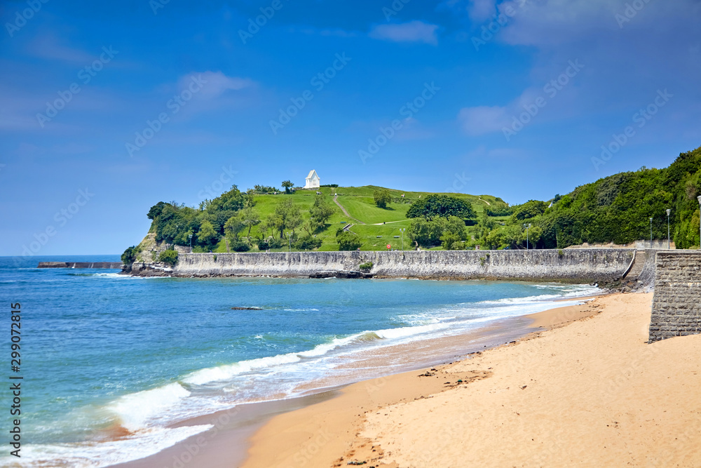 Landscape with a sandy beach and embankment of Saint-Jean-de-Luz, green hill with white chapel on top (Basque Country, Atlantic coast, France). Coastal french town at sunny summer day. Sea shore