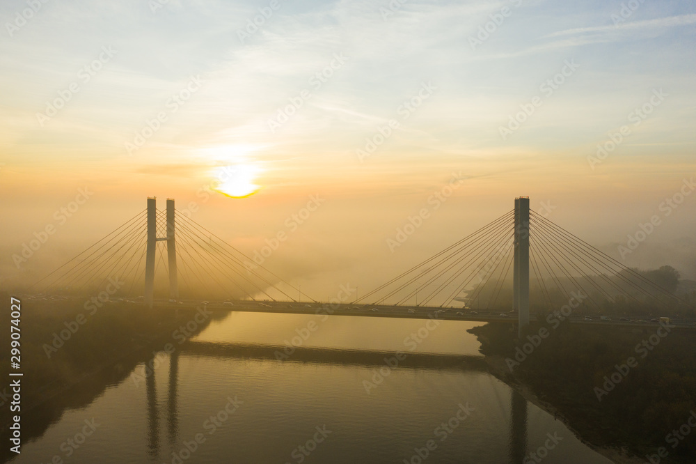 Obraz premium Aerial view of a bridge with cars in the fog. Warsaw. Poland. Drone shot at the traffic of a vehicle traveling in traffic jam on a bridge over a river. Drone shot into the fog at sunrise over a bridg