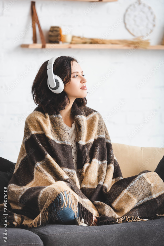 beautiful girl thinking and listening music with headphones while sitting in blanket - obrazy, fototapety, plakaty 