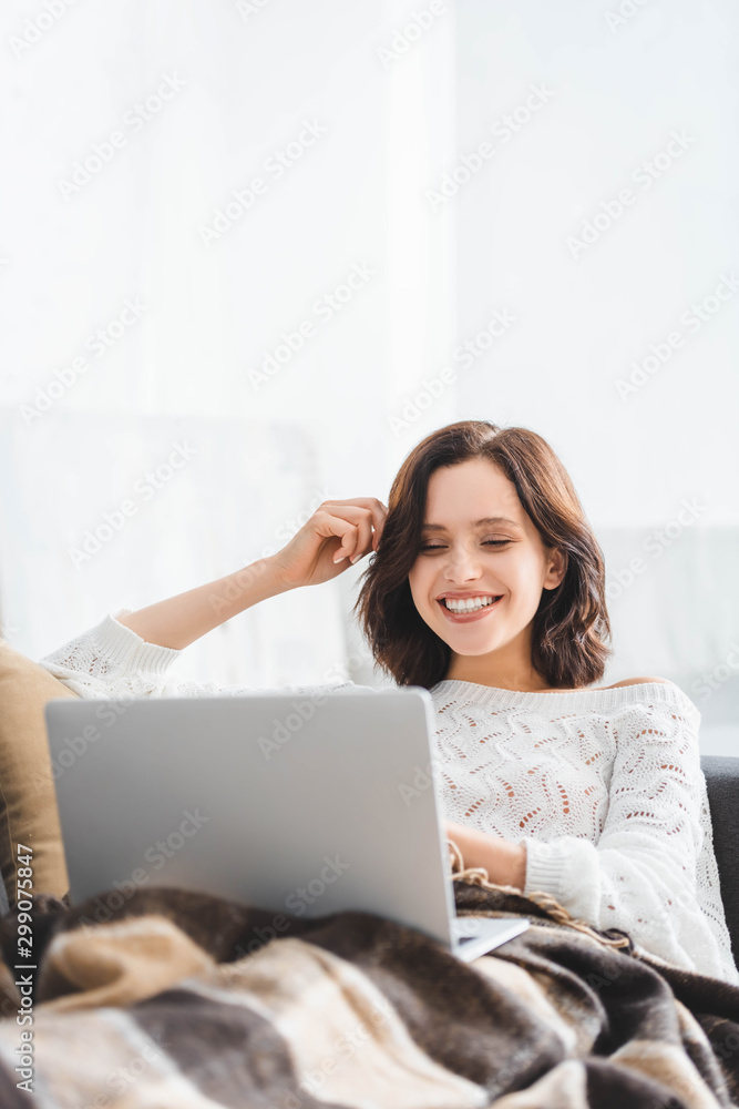 cheerful woman in blanket using laptop on sofa in cozy living room cheerful woman in blanket using laptop on sofa in cozy living room
