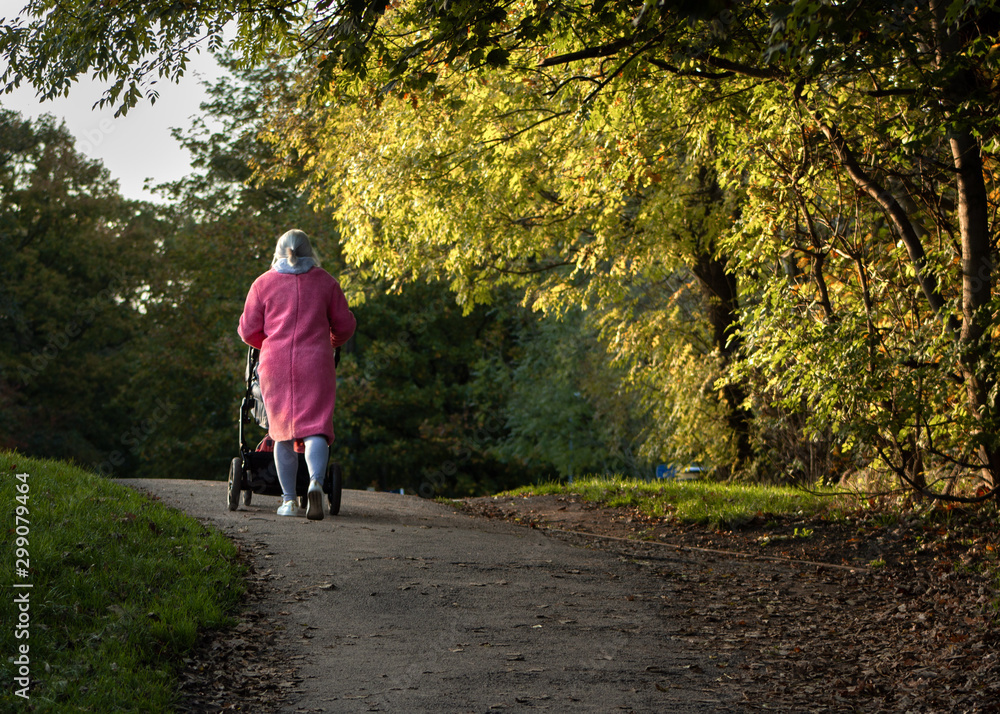 Fototapeta premium Despite the autumn sunshine a woman wraps up warm in a pink coat when taking a child in a pushchair on the tow path of the Leeds & Liverpool Canal at Hirst Wood