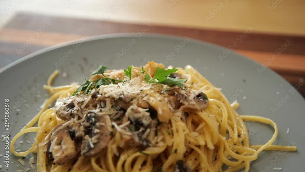 Close up rotating side view of linguine pasta topped with mushrooms, parmesan cheese, and parsley on a gray plate on a striped, wooden cutting board. Close up rotating side view of linguine pasta topped with mushrooms, parmesan cheese, and parsley on a gray plate on a striped, wooden cutting board.