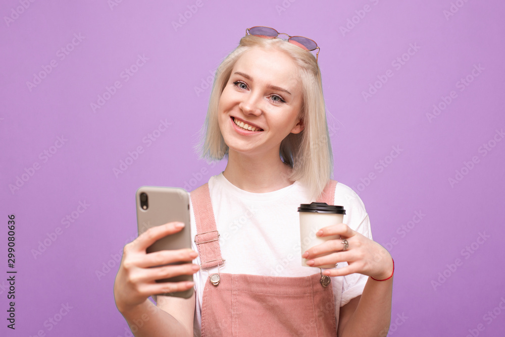 Portrait of a happy girl standing on a purple background with a smartphone and a cup of coffee in hand, looking into the camera and smiling, wearing a cute casual wear. Isolated. Portrait of a happy girl standing on a purple background with a smartphone and a cup of coffee in hand, looking into the camera and smiling, wearing a cute casual wear. Isolated.