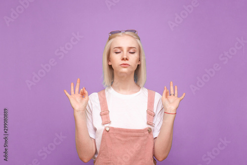 Sweet girl in a bright casual dress mediates with her eyes closed, standing on a purple background. Lady is a teenager with light hair doing meditation, isolated photo