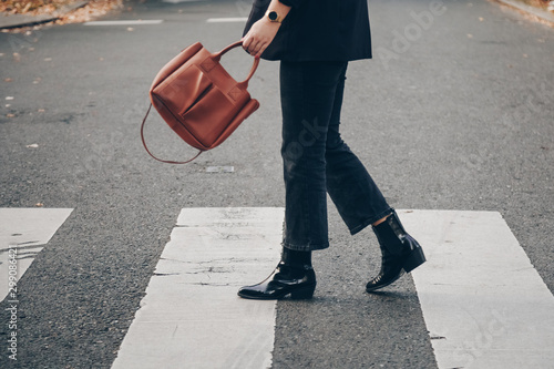 fashion blogger autumn 2019 outfit details. fashionable woman wearing black jeans, black ankle shoes a brown tan trendy leather handbag. crossing the street. detail of a perfect fall fashion outfit.  photo