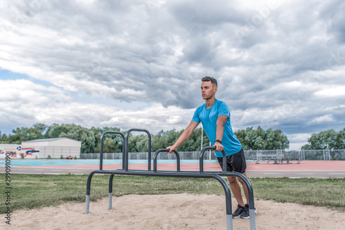 Athletic man exercises push-ups from horizontal bar, summer afternoon training in city, active lifestyle, modern fitness workout. Free space motivation text. Background clouds sports field.