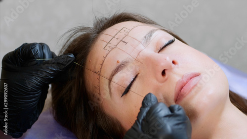 The process of shaping eyebrows by a beauty master with a thread for a young lady in the studio before the microblading. Close up view of a face while the procedure of marking lines.