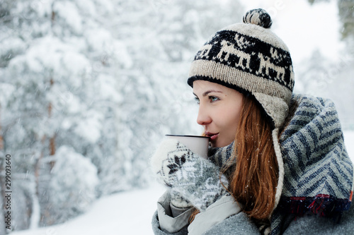 Beautiful young woman standing among snowy trees in winter forest and enjoying cup of hot tea.