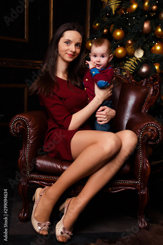 Mom sits on a chair with a baby in her arms, hugs and kisses. Christmas gift, family and happiness, Christmas tree and holiday