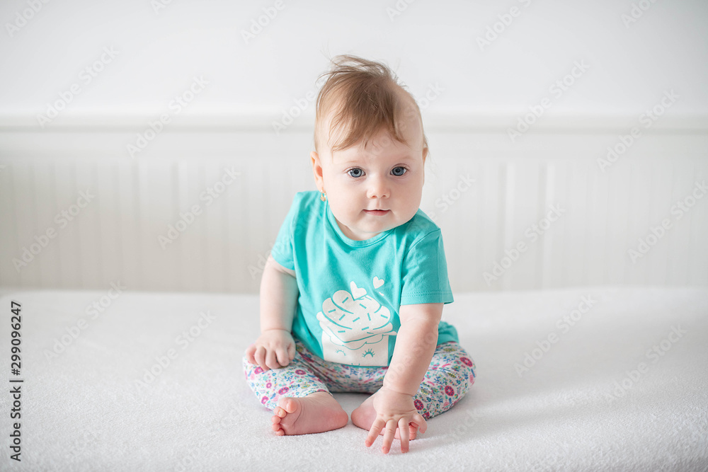 Fototapeta premium Portrait of adorable Caucasian baby girl with blue eyes, looking at the camera calmly, with curiosity, interactivity or inquisitiveness and sitting in a pristine white baby cot while holding her toes