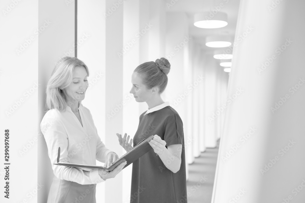 Fototapeta premium Black and white photo of Businesswomen discussing over document while standing in corridor at office