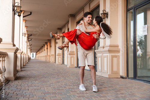 Fototapeta Image of young unshaven man carrying happy woman in hands and smiling