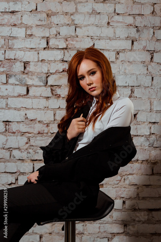 A young elegant red-haired lady in a black suit with jacket and trousers sits on a chair and looks seriously. Portrait of a girl on the white brick background.  © Dzmitry