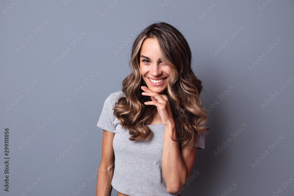 Fototapeta premium Atrakcyjna kobieta z długimi kręconymi włosami jest bardzo zadowolona, uśmiechając się z szerokim uśmiechem pokazując jej idealne zęby w pomieszczeniu.