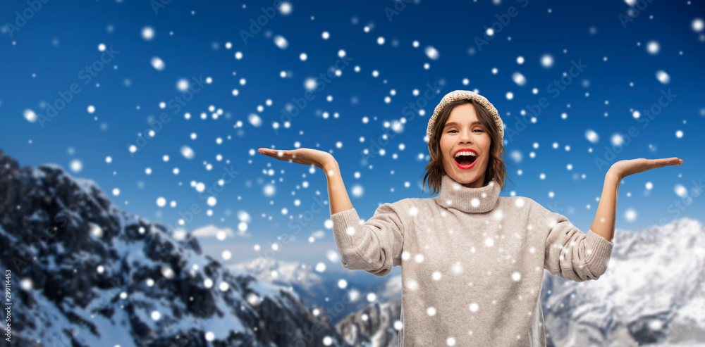christmas, season and people concept - happy smiling young woman in knitted winter hat and sweater holding something on empty hand palm over snow and alps mountains background
