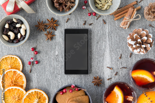 christmas, winter holidays and technology concept - smartphone, hot mulled wine, dry orange slices and aromatic spices on grey background