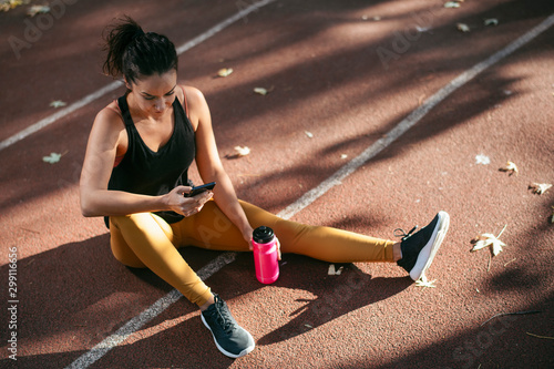 Young woman working out. Beautiful athlete woman sitting on running track after jogging and using phone.