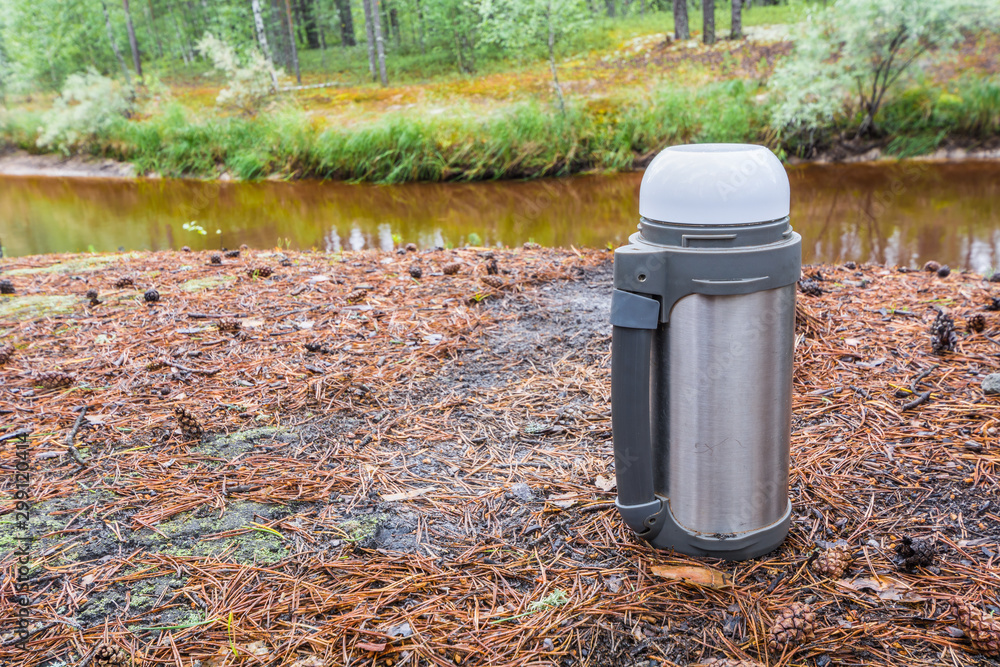 Hot tea thermos stands on ground with pine needles in forest against taiga river. Hot tea thermos stands on ground with pine needles in forest against taiga river.