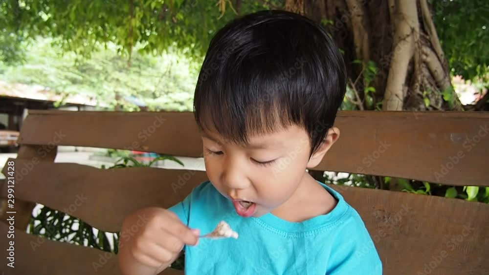 Asian child boy in blue T-shirt sitting and eating ice cream by spoon with happy relax face outdoor with natural background. Kid enjoy cold dessert food in summer happy day. Asian child boy in blue T-shirt sitting and eating ice cream by spoon with happy relax face outdoor with natural background. Kid enjoy cold dessert food in summer happy day.