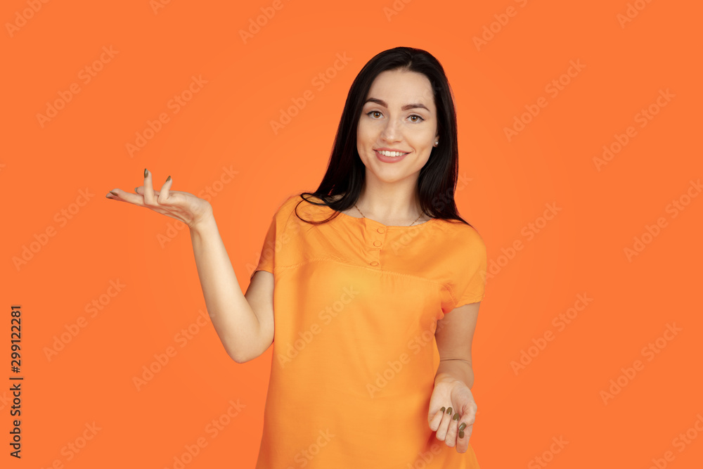 Caucasian young woman's portrait on orange studio background. Beautiful female brunette model in shirt. Concept of human emotions, facial expression, sales, ad. Copyspace. Pointing, showing, smiling. Caucasian young woman's portrait on orange studio background. Beautiful female brunette model in shirt. Concept of human emotions, facial expression, sales, ad. Copyspace. Pointing, showing, smiling.