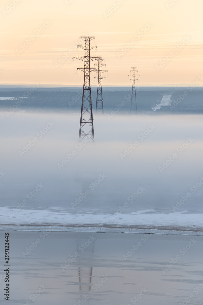 Power lines standing on the Lena River, Yakutia, Republic of Sakha, Russia. Dense fog over the river at gold sunrise. Beginning of winter. Winter landscape with river and fog. Foggy morning