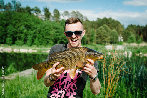 Happy cheerful young fisherman hold a big fish carp on a background of lake and nature. Fishing background. Good catch. Trophy fish. angler.