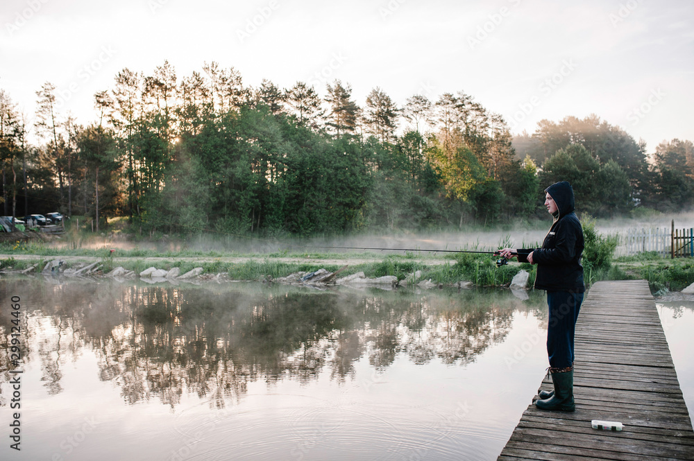 Fisherman with fishing rod on bridge. Sunrise. Fishing for pike, perch, carp. Fog, grass, trees against the backdrop of lakes, nature. Fishing background. Misty morning. the wild nature. river
