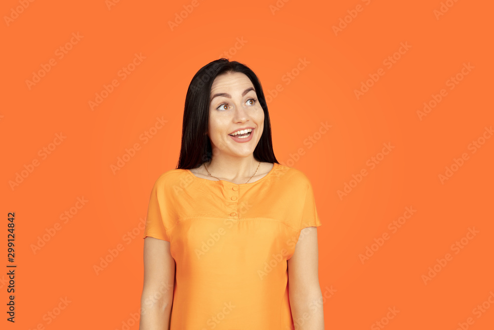 Obraz premium Caucasian young woman's portrait on orange studio background. Beautiful female brunette model in shirt. Concept of human emotions, facial expression, sales, ad. Copyspace. Looks dreamful, smiling.