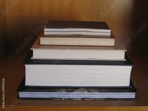 Libros apilados encima uno de otro en forma de escalera, atrio, distintos tamaños y pastas en rincón de estantería para despacho y trabajo, luz de penumbra, el último con tapa ligeramente levantada 