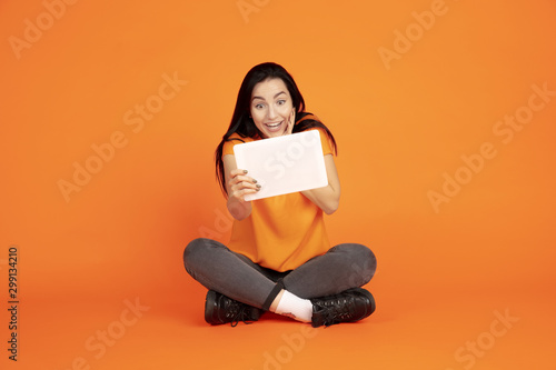 Caucasian young woman's portrait on orange studio background. Beautiful female brunette model in shirt. Concept of human emotions, facial expression, sales, ad. Copyspace. Using tablet, vlogging.