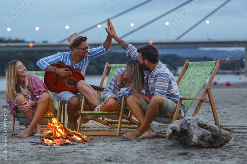 Group of friends sitting around camp fire at the beach at the autumn evening.They play guitar and singing.