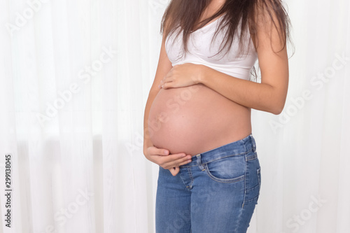 Pregnant woman in jeans and white top holds hands on belly on a white background. Pregnancy, maternity, preparation and expectation concept. Beautiful tender mood photo of pregnancy. Free Download Pregnant woman in jeans and white top holds hands on belly on a white background. Pregnancy, maternity, preparation and expectation concept. Beautiful tender mood photo of pregnancy. - GFXLoad